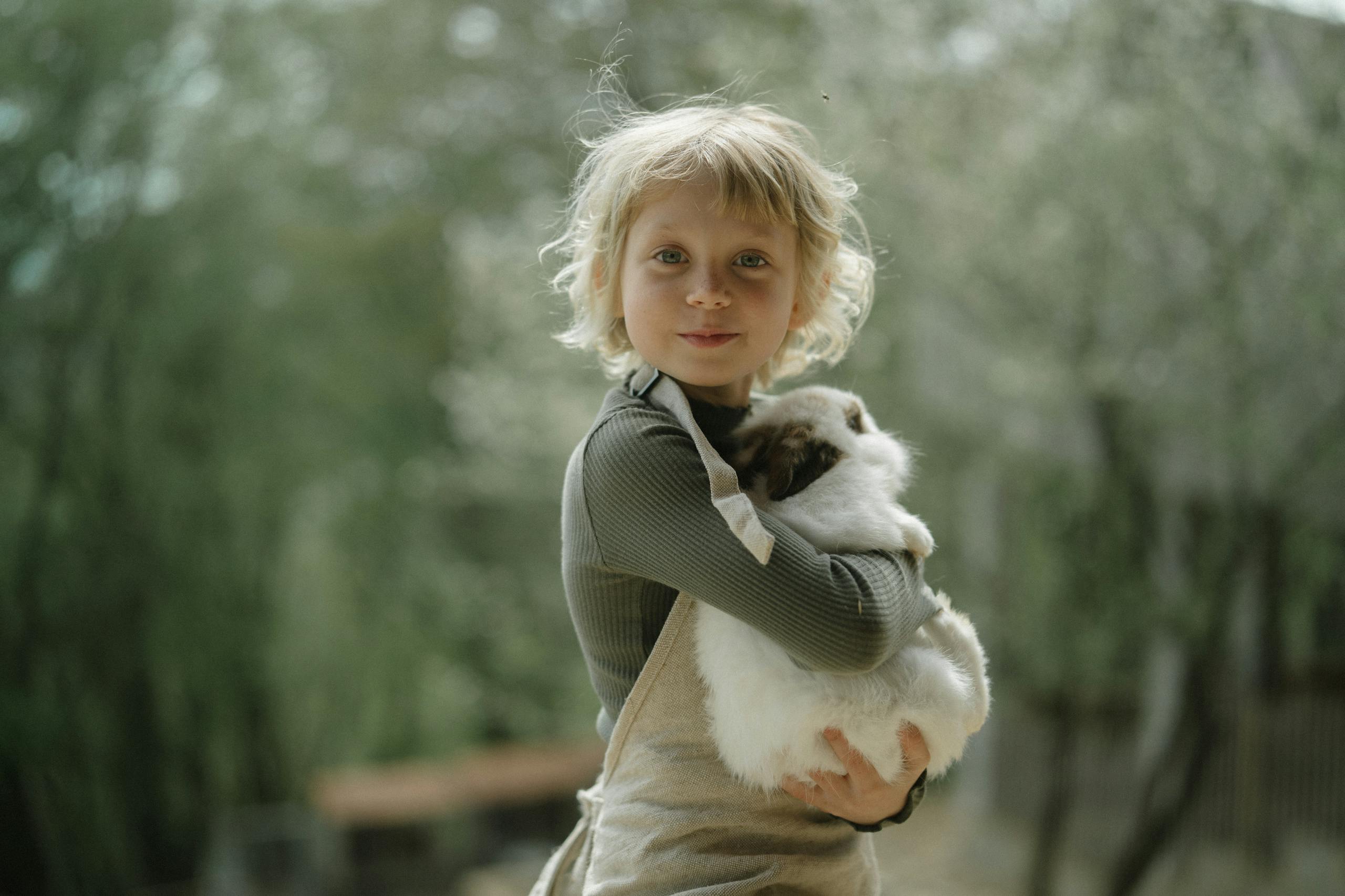A cute blonde child smiling while hugging a fluffy rabbit in an outdoor setting with a blurred background.