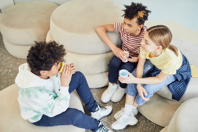 From above of group of multiethnic children sitting on soft pillows and drinking hot beverage while spending time together in living room