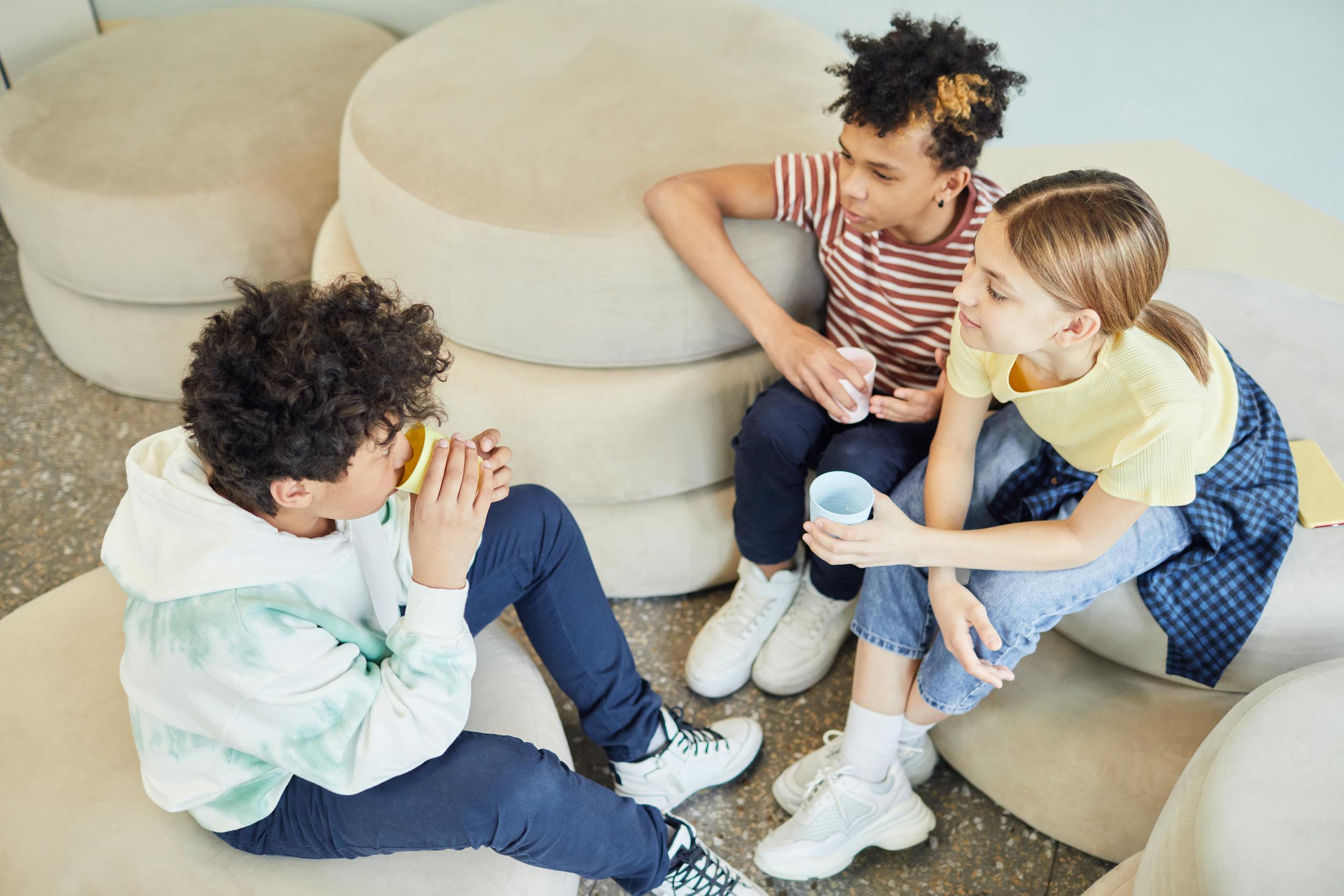 From above of group of multiethnic children sitting on soft pillows and drinking hot beverage while spending time together in living room
