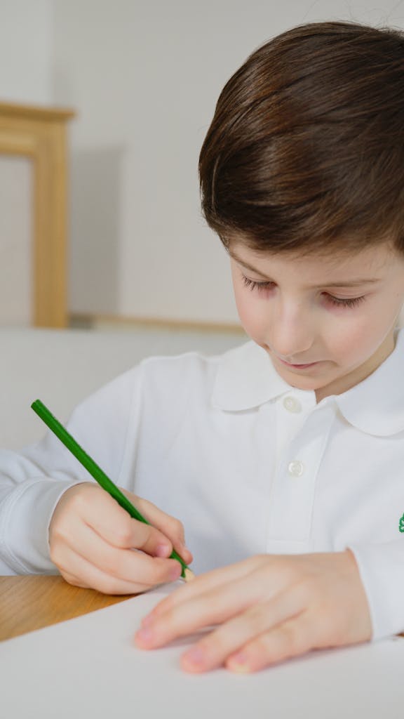 Caucasian child in white shirt writing attentively with a green pencil indoors.