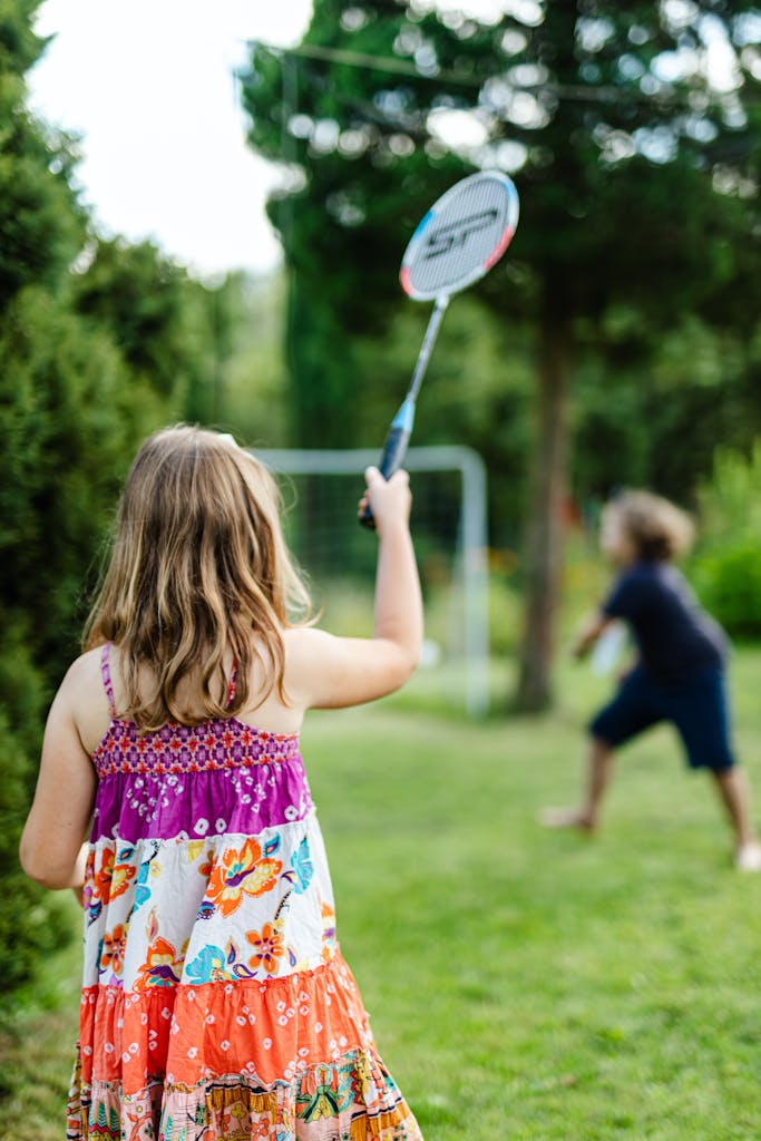 Two children enjoy a game of badminton outdoors in a sunny park setting.