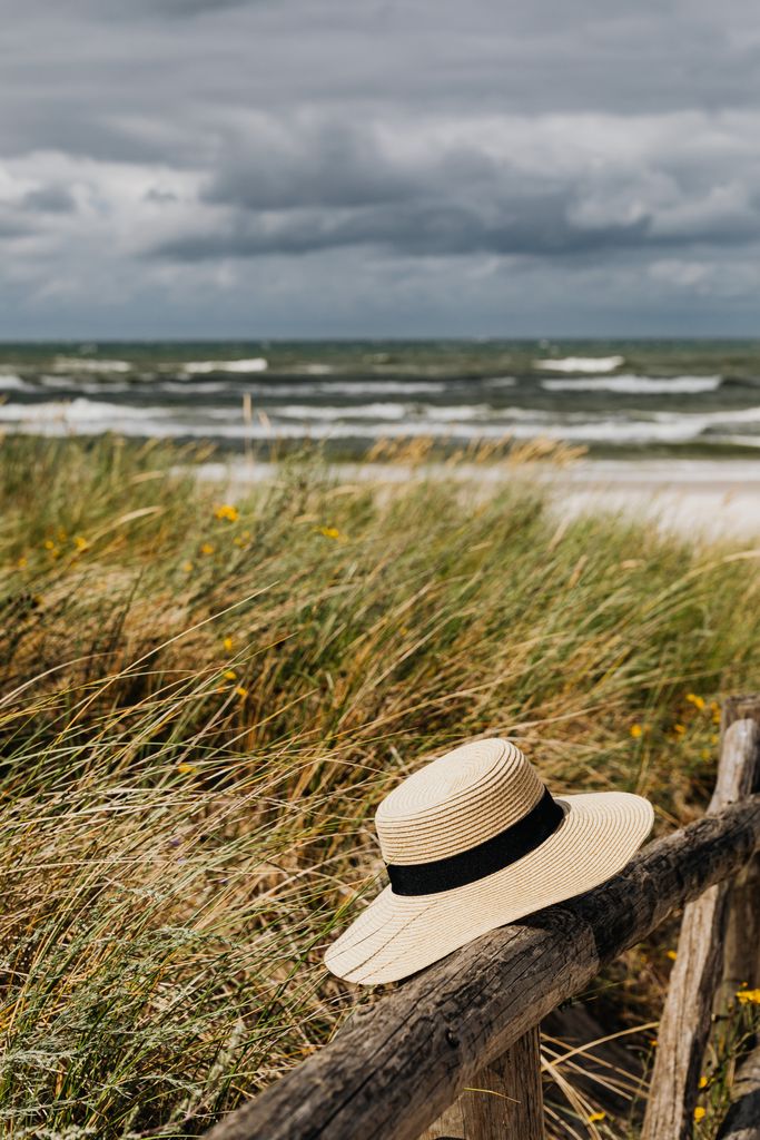 A straw hat rests on a wooden fence against a backdrop of grassy dunes and ocean waves.
