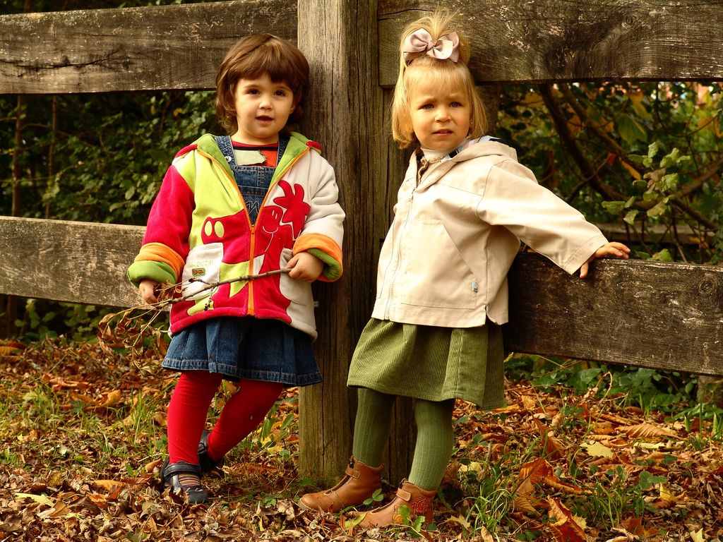 Two young children dressed warmly pose by a wooden fence during autumn.